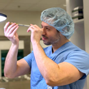 A healthcare worker in blue scrubs and a hair cap prepares a syringe, holding it up to eye level under bright light in a medical setting.