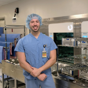 A smiling medical professional in blue scrubs and a hairnet stands in a clean, stainless steel hospital sterilization or surgical prep area with equipment and trays visible in the background.