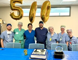 Seven smiling medical staff in scrubs stand behind a blue table with a decorated cake and a trophy. Large gold balloons form the number 500, celebrating a milestone event in a brightly lit room.