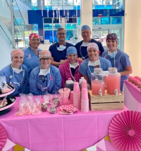 A group of smiling healthcare workers in scrubs and pink accessories stand behind a pink-themed table with lemonade, cupcakes, and treats, inside a bright, modern building with blue accents.