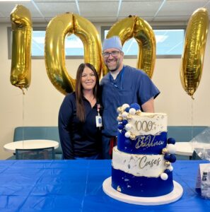 Two people stand smiling behind a blue and white cake that says “1000 DaVinci Cases,” with large gold balloon numbers “1000” in the background, celebrating a milestone in a decorated room.