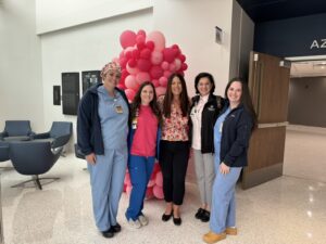 Five women, some in medical scrubs and some in business attire, stand smiling together in a bright lobby area. Behind them is a large pink balloon arrangement.