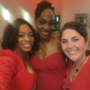 Three women smiling, wearing elegant red dresses, pose closely together for a photo in a warmly lit indoor setting.