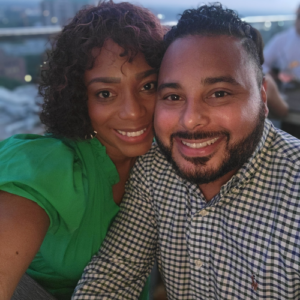 A smiling couple poses for a close-up selfie outdoors. The woman wears a green top, and the man wears a checkered shirt. They appear happy, with blurred city lights and people in the background.