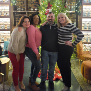 Four adults stand smiling in front of a decorated Christmas tree. Three women and one man have their arms around each other in a festive, warmly lit indoor setting.