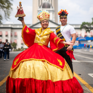 A woman in a vibrant red and gold traditional dress, holding a religious statue, stands in front of a man wearing a feathered headdress and sash during a festive outdoor celebration. A large building is visible in the background.