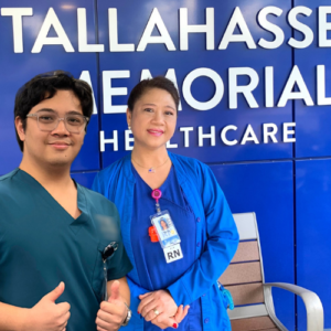 Two healthcare professionals stand in front of a blue wall reading "Tallahassee Memorial Healthcare." One wears green scrubs and glasses, while the other, wearing blue scrubs and an RN badge, smiles at the camera.