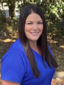 A woman with long straight brown hair, wearing a bright blue top, smiles while standing outdoors with leafy green trees and sunlight in the background.