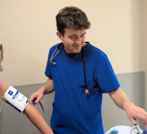 A healthcare worker in blue scrubs and a stethoscope checks a patient's blood pressure, looking at a monitor while holding the patient's arm with a cuff attached.
