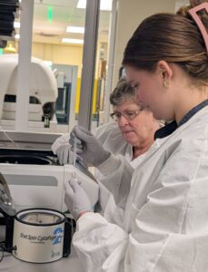 Two women in lab coats and gloves work together in a laboratory. One is holding a pipette and tube, while the other observes closely. A centrifuge labeled "StatSpin CytoFuge" is visible on the counter nearby.