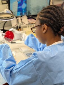 A person wearing a blue lab coat and gloves examines a petri dish with red agar in a laboratory, sitting at a desk with scientific equipment and a computer screen in the background.