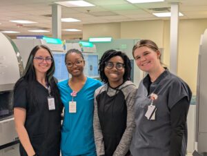 Four women stand together smiling in a laboratory, wearing scrubs or work uniforms with ID badges. Laboratory machines and equipment are visible in the background.