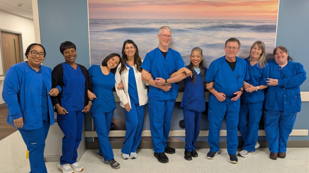 Eight smiling healthcare workers stand together in blue scrubs, arms linked, in a hospital hallway with a large beach scene photo on the wall behind them. The mood is friendly and positive.