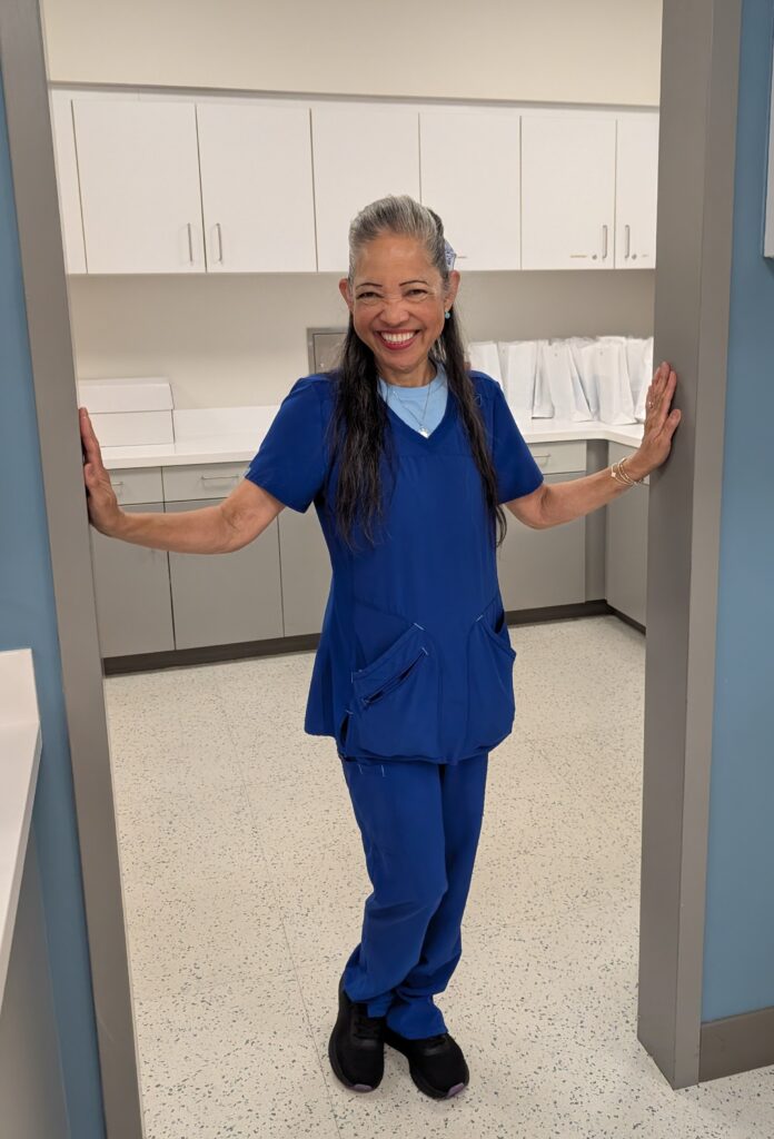 A smiling woman wearing blue medical scrubs and black shoes stands in a doorway with her arms outstretched, in a clean, modern medical office with white cabinets in the background.