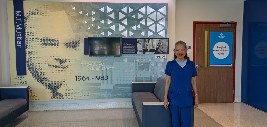 A smiling healthcare worker in blue scrubs stands in a hospital waiting area near a wall display honoring M.T. Mustian, with a sign for the Surgical Pre-Admission Clinic visible on a nearby door.