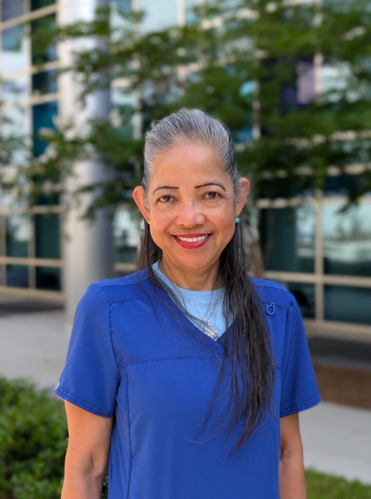 A woman with long gray hair smiles while standing outside in front of a building with glass windows. She is wearing a blue scrub top and a light blue shirt underneath. Greenery is visible in the background.